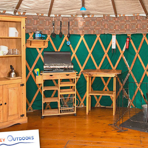 Yurt interior with wooden furniture and equipment, featuring Anglesey Outdoors branding.