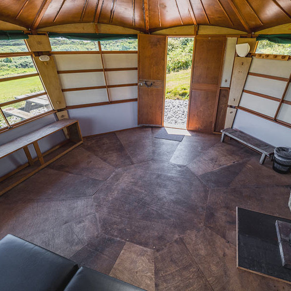 Interior of a yurt with wooden walls and floor, large windows, and outdoor scenery.